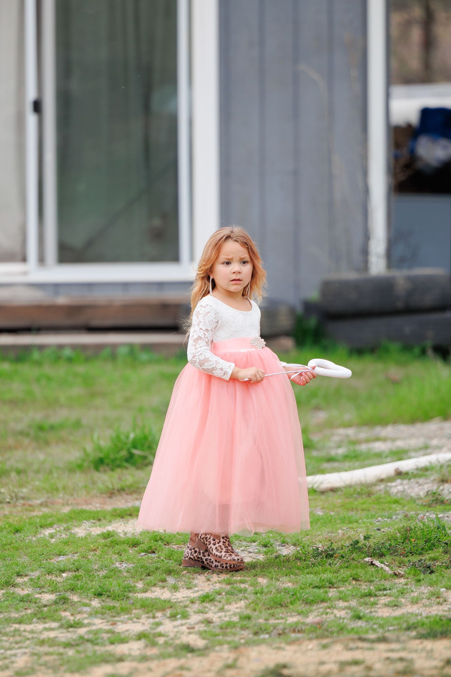 A young child wearing a pink tulle dress and leopard print boots stands on grass outside a building.