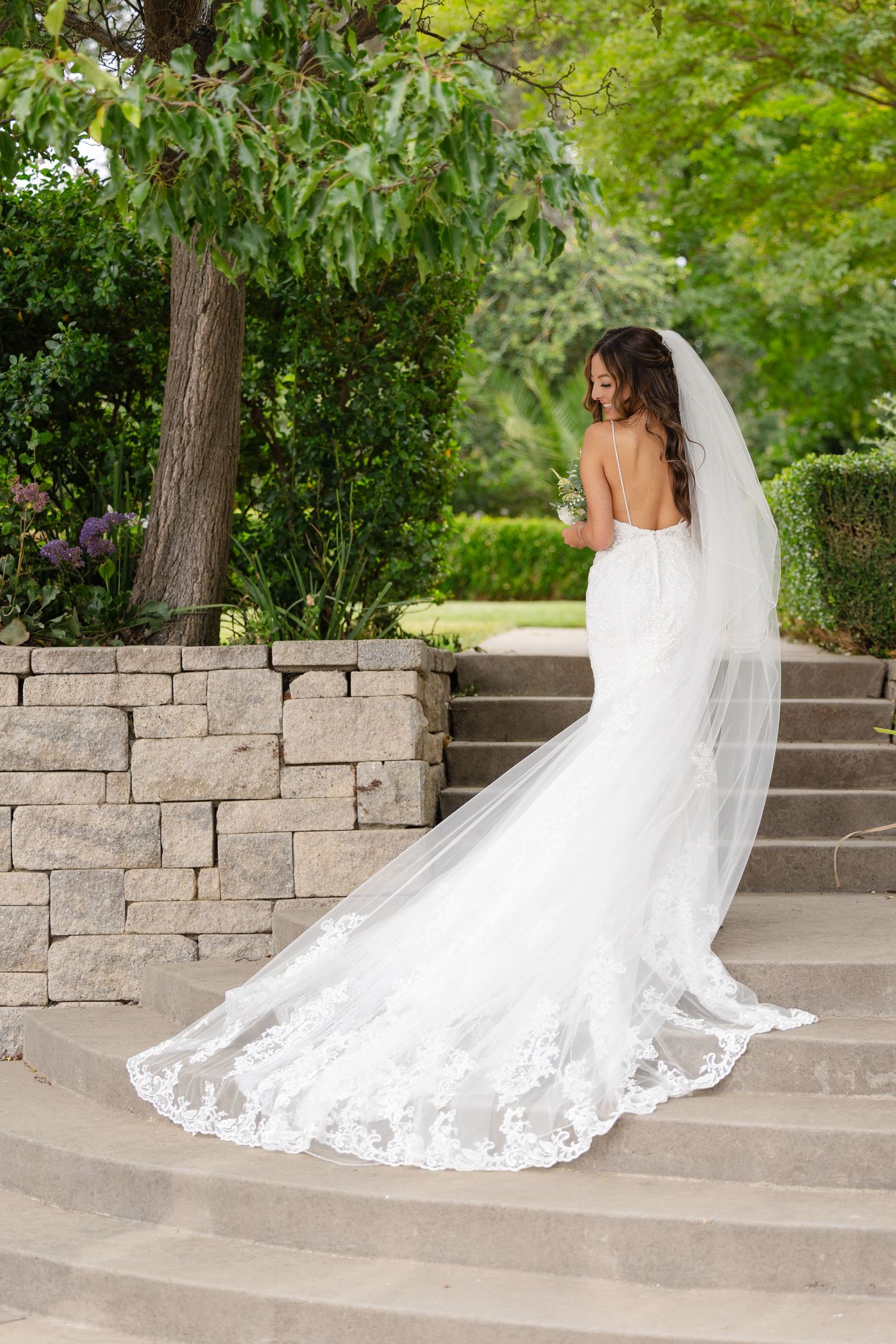 A stunning white lace wedding gown with a long flowing cathedral veil cascades down stone steps in a garden setting.