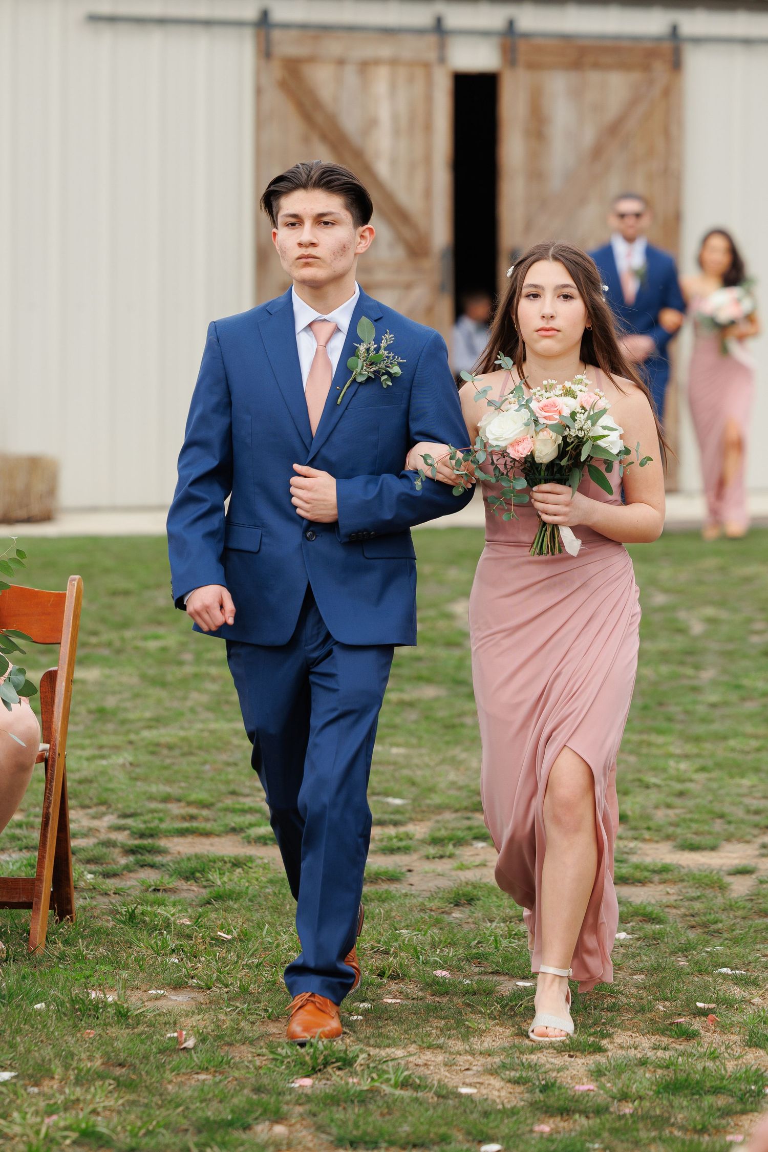 Wedding party members in navy suits and dusty rose dresses walk together on a grassy lawn during outdoor ceremony.