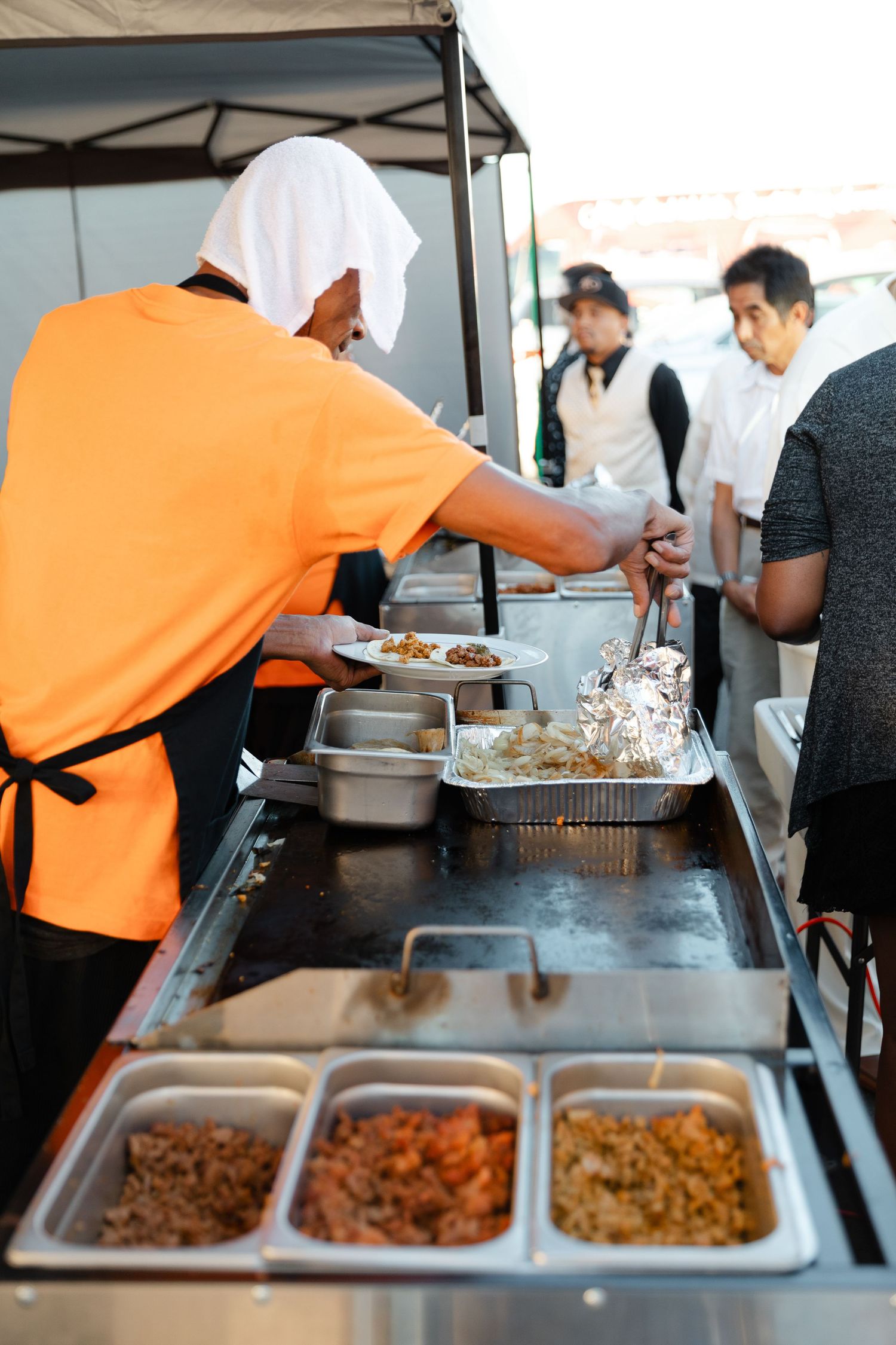 Food service workers in orange shirts prepare and serve food at a buffet line with multiple stainless steel serving trays.
