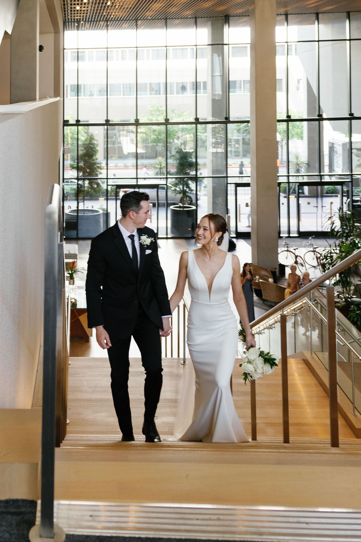 Elegant wedding couple walks through a modern lobby with floor-to-ceiling windows and sleek architectural details.