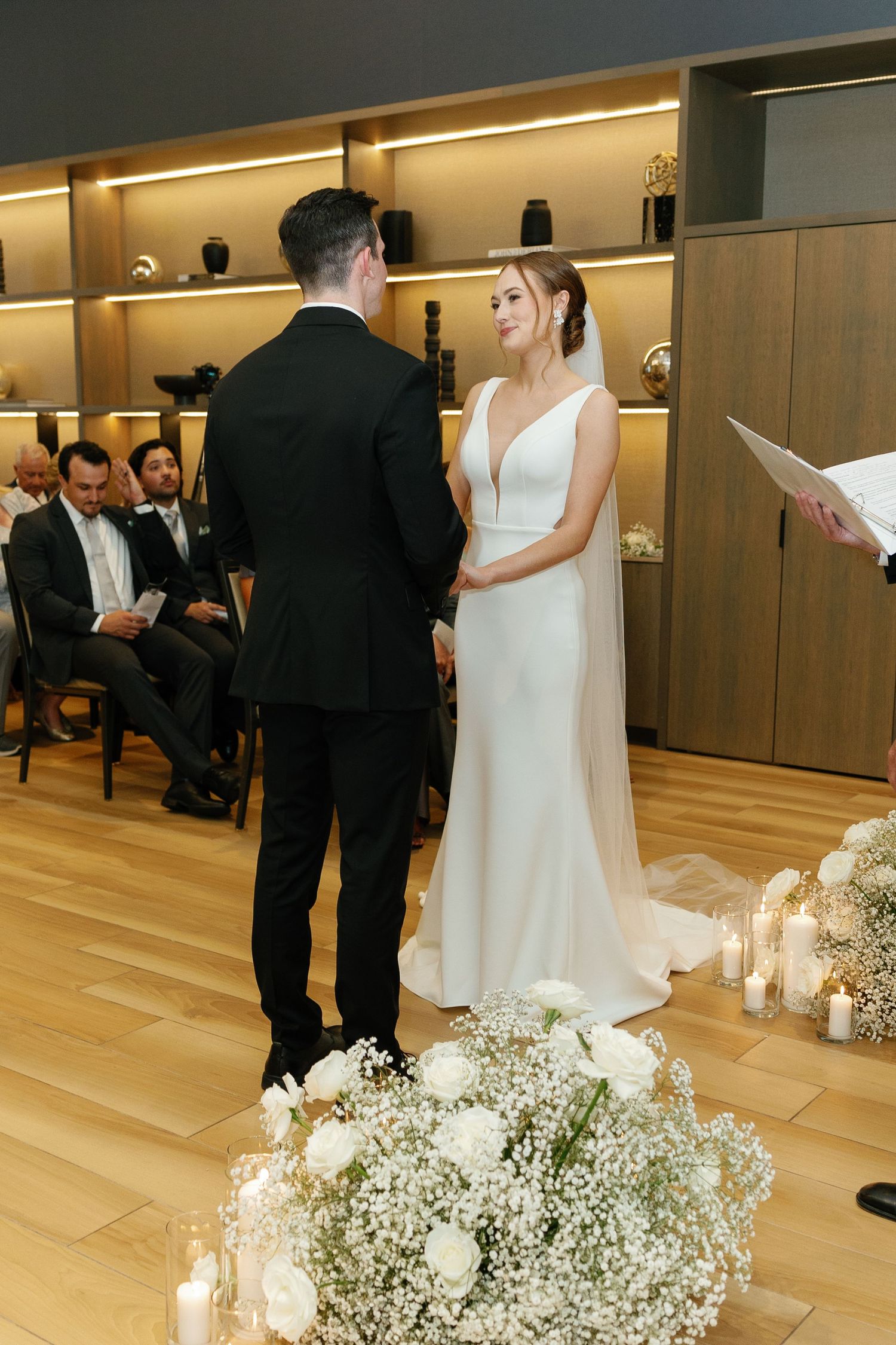 A wedding ceremony in a modern venue with illuminated wooden shelving and delicate white floral arrangements.