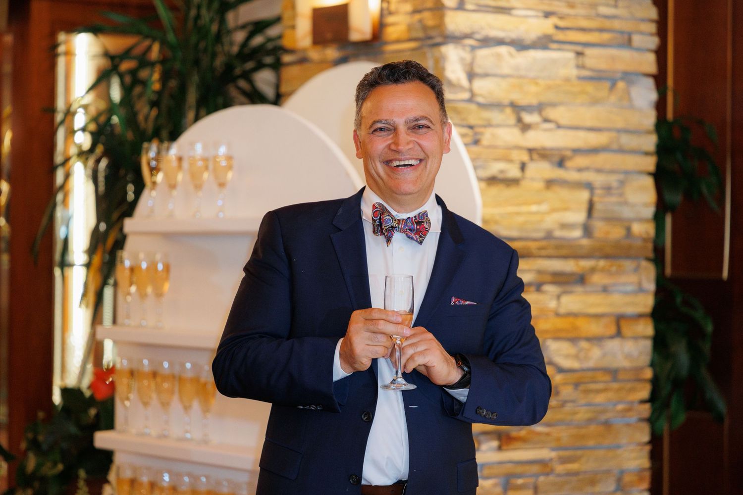 A person in a navy suit and bow tie sits at a formal event with stone walls and warm lighting in the background.