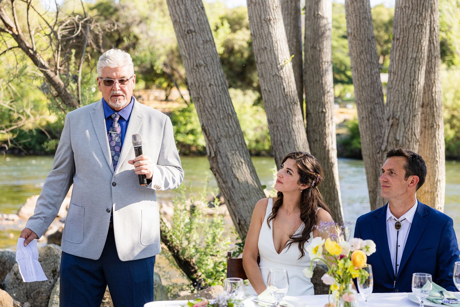 Wedding party laughs together during outdoor reception under tall pine trees near a lake.