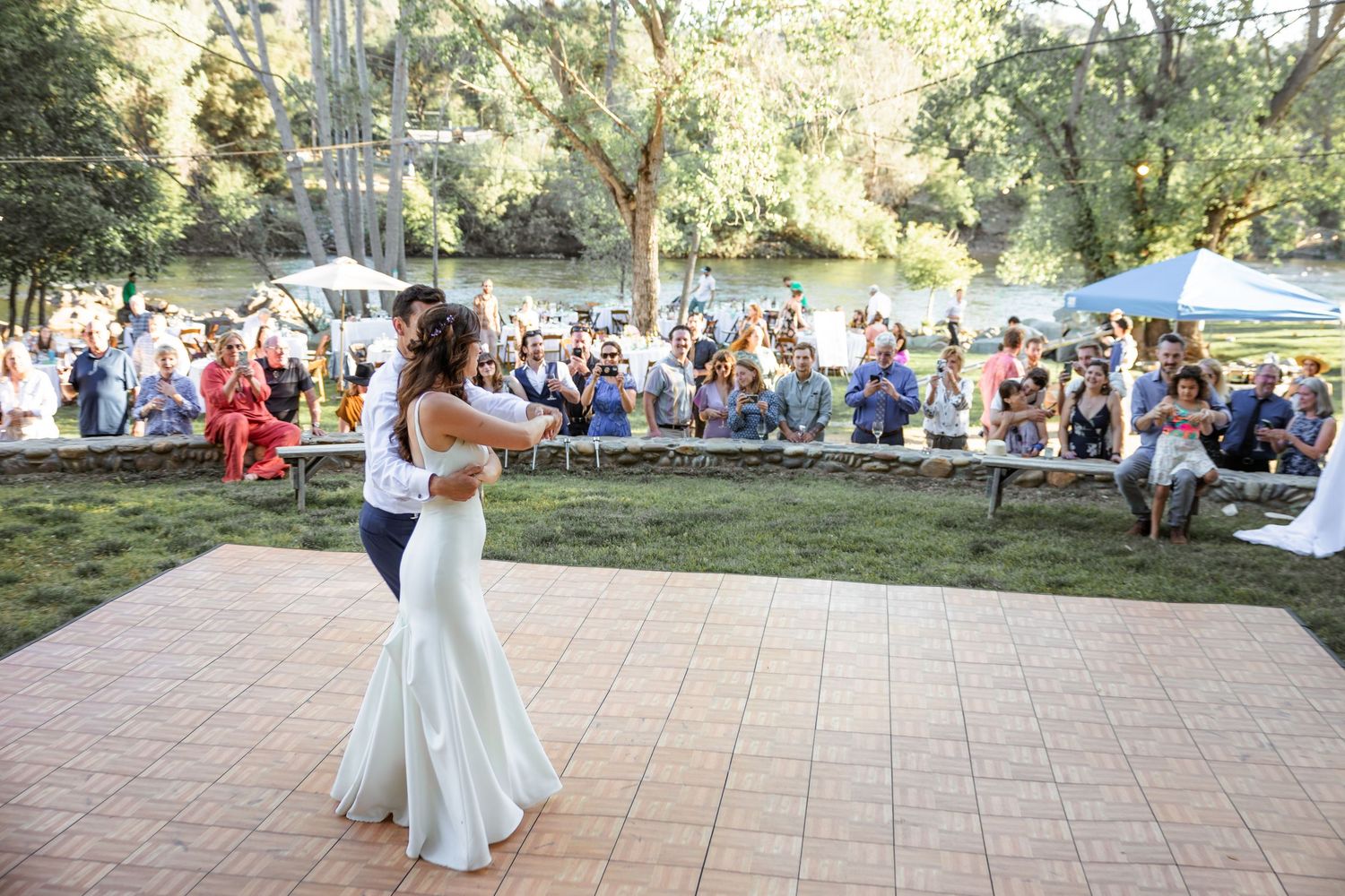 A couple performs their first dance at an outdoor wedding reception on a stone patio surrounded by guests.