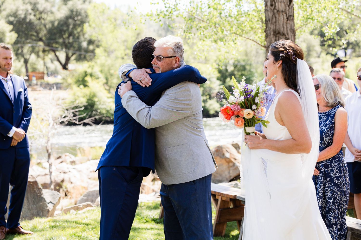 Two people embrace warmly at an outdoor wedding ceremony while a bride in white holds her bouquet nearby.