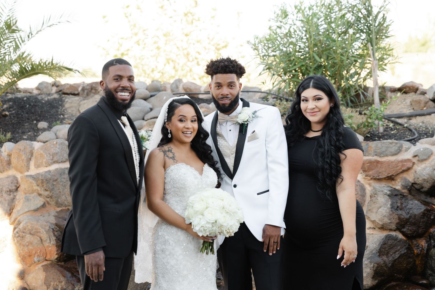 A wedding party poses together outdoors against a stone wall backdrop with greenery and natural lighting.