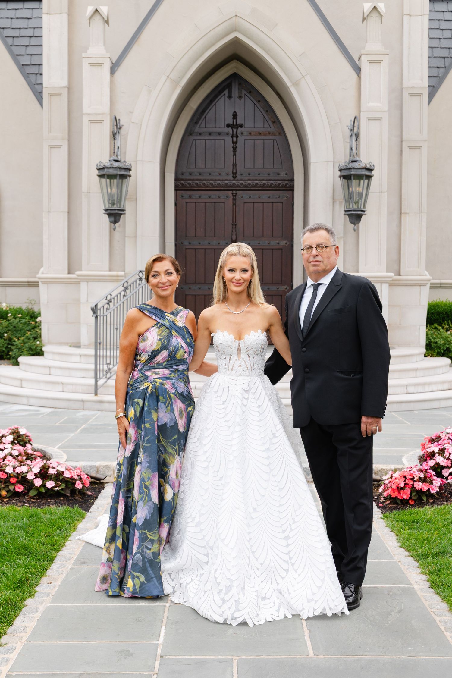 A wedding party poses outside a Gothic-style church entrance with white stone arches and pink flowers.