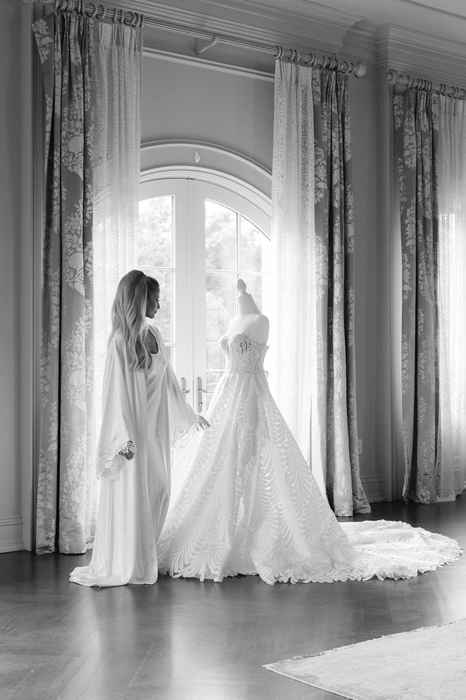 A collection of elegant white wedding dresses displayed in a bright room with arched windows and flowing curtains.
