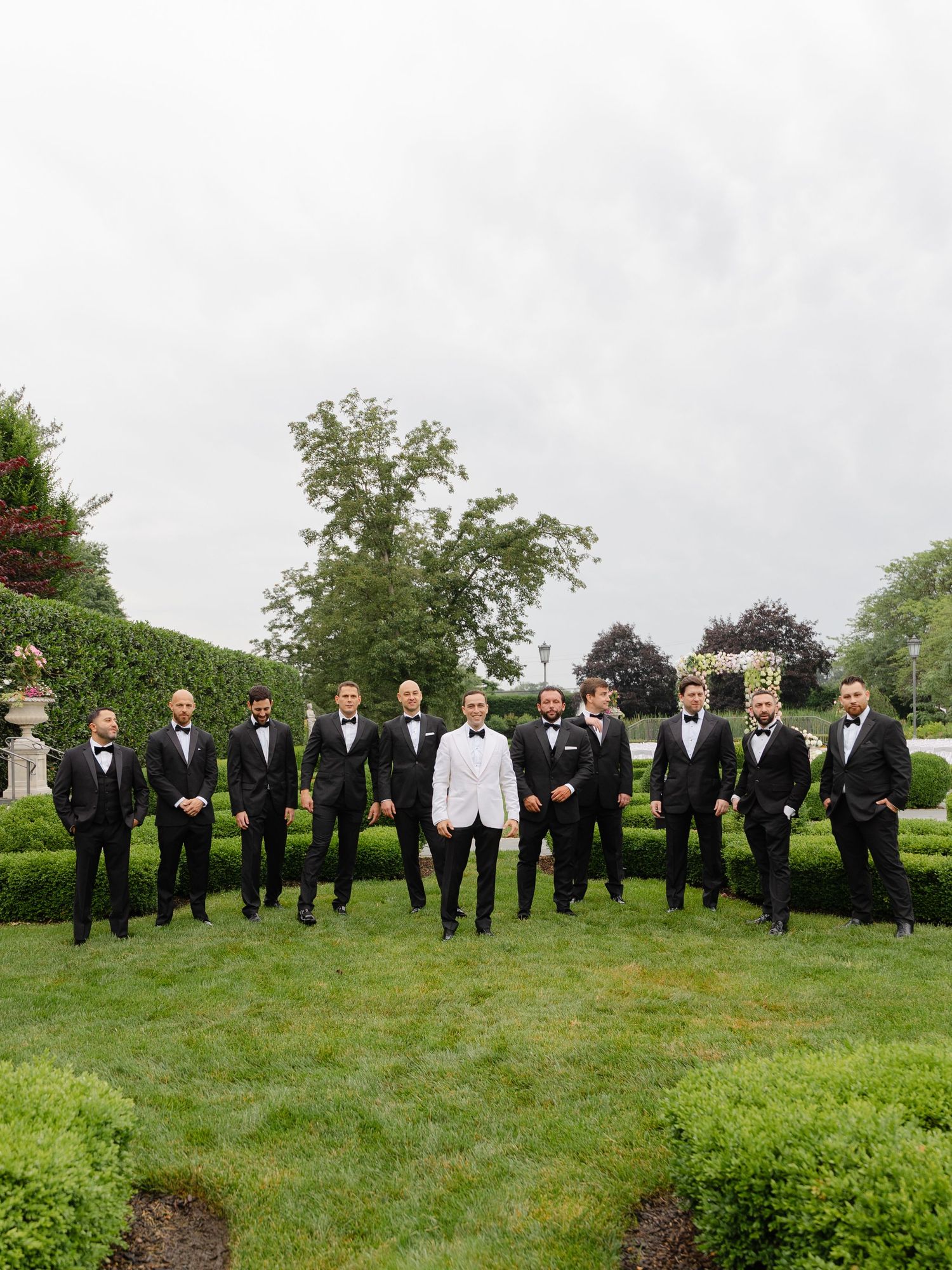 A wedding party in formal attire poses on a manicured lawn with hedges and garden statues in the background.