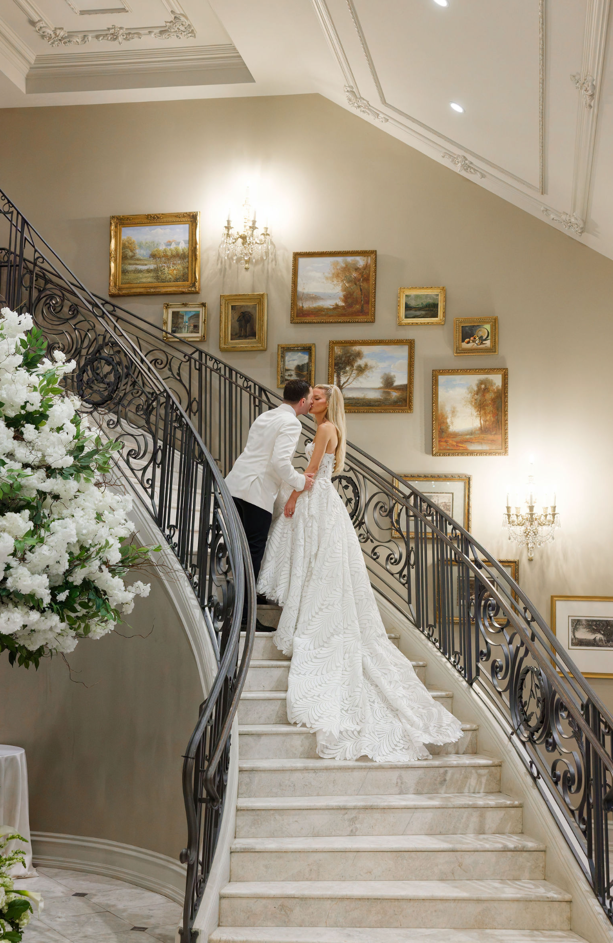 Elegant wedding couple on grand marble staircase with ornate iron railing and gold-framed artwork adorning the walls.
