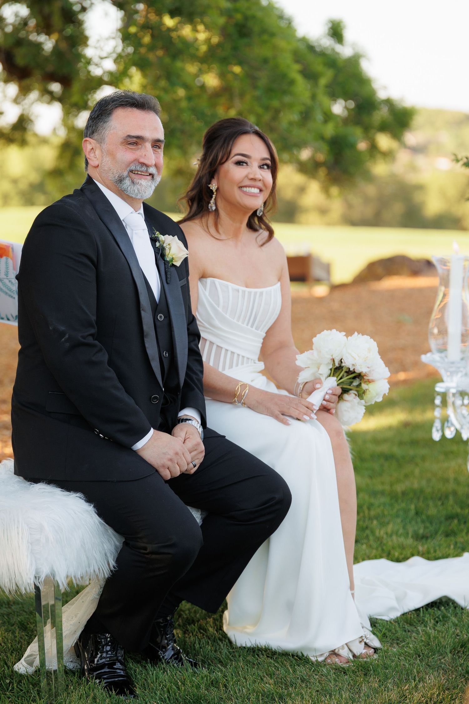 A couple in wedding attire sits together on grass during an outdoor photoshoot at sunset.