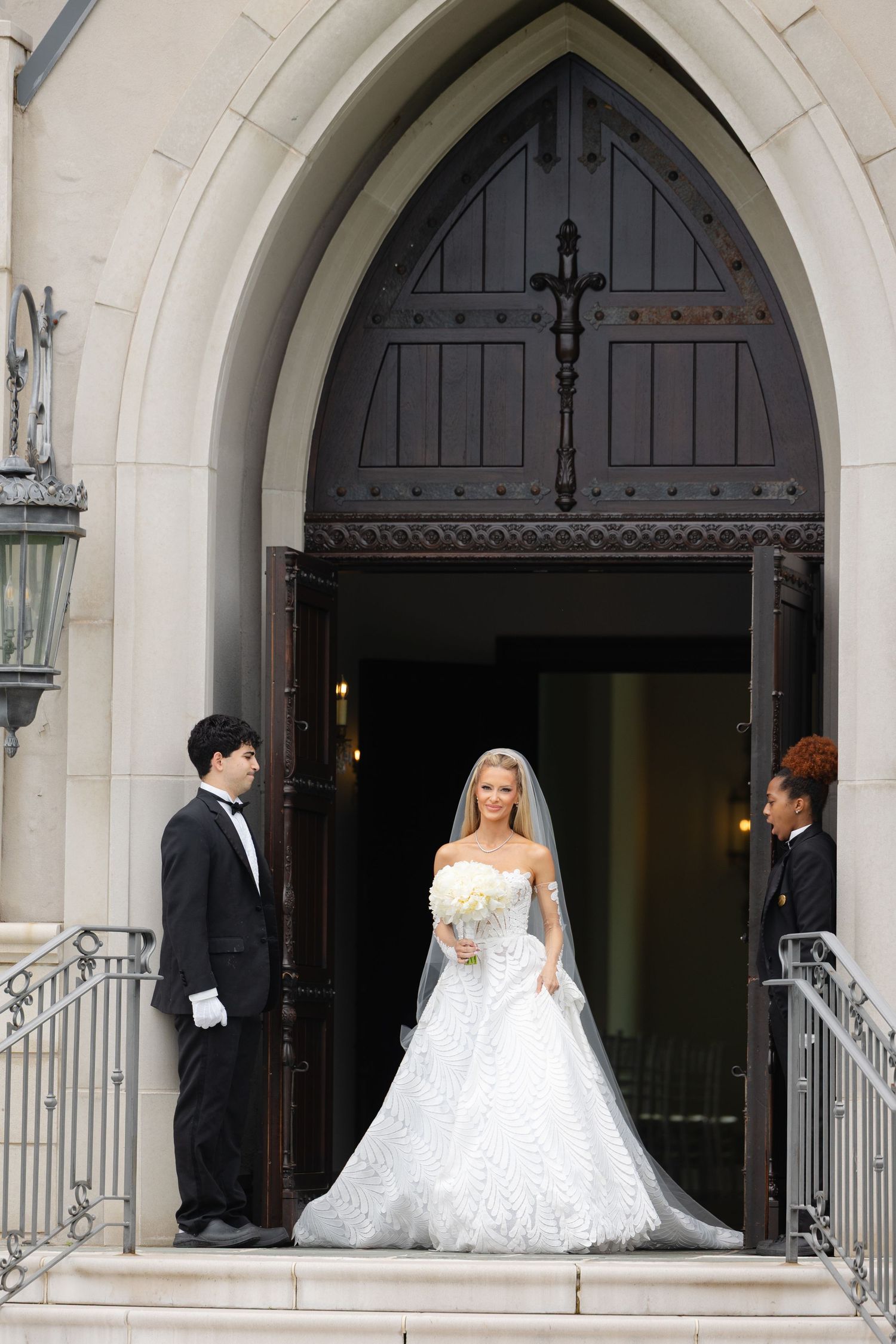 Bride in white gown holding bouquet exits church doors, flanked by two attendants in formal attire.