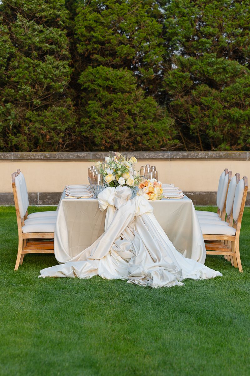 Romantic outdoor table setting with draped white linens and floral centerpiece against a garden wall backdrop.
