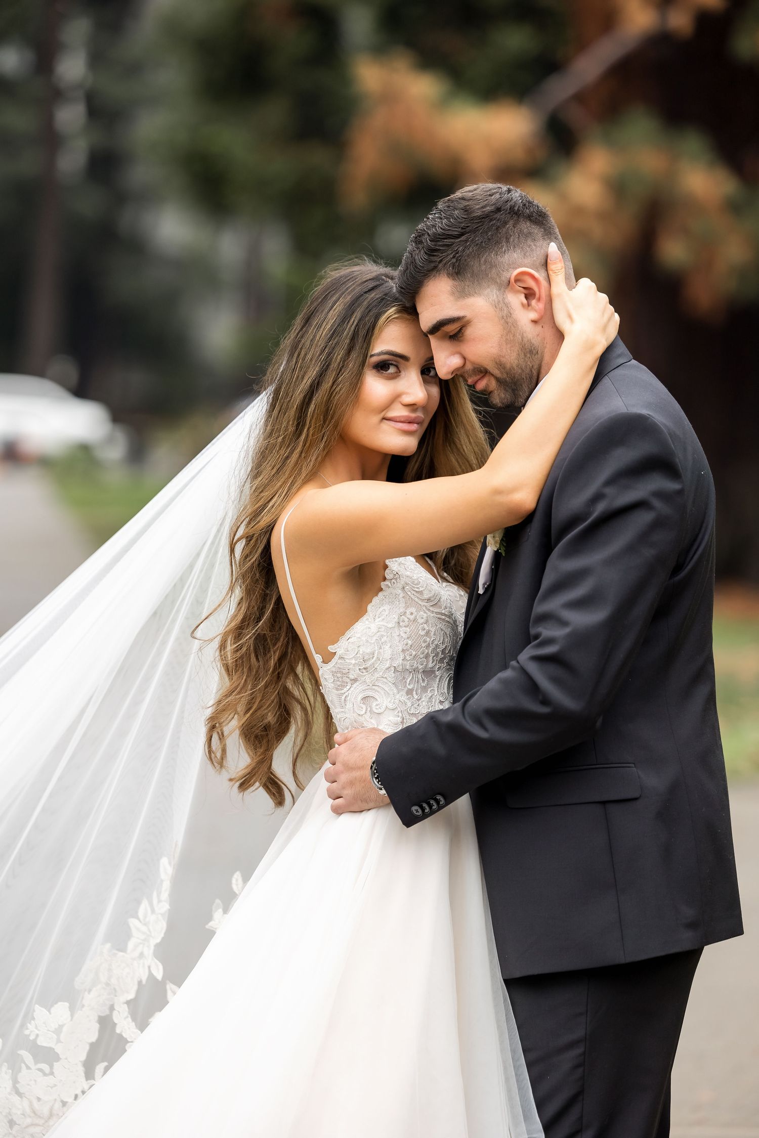 Romantic wedding couple embraces outdoors while bride's long veil flows in the breeze.