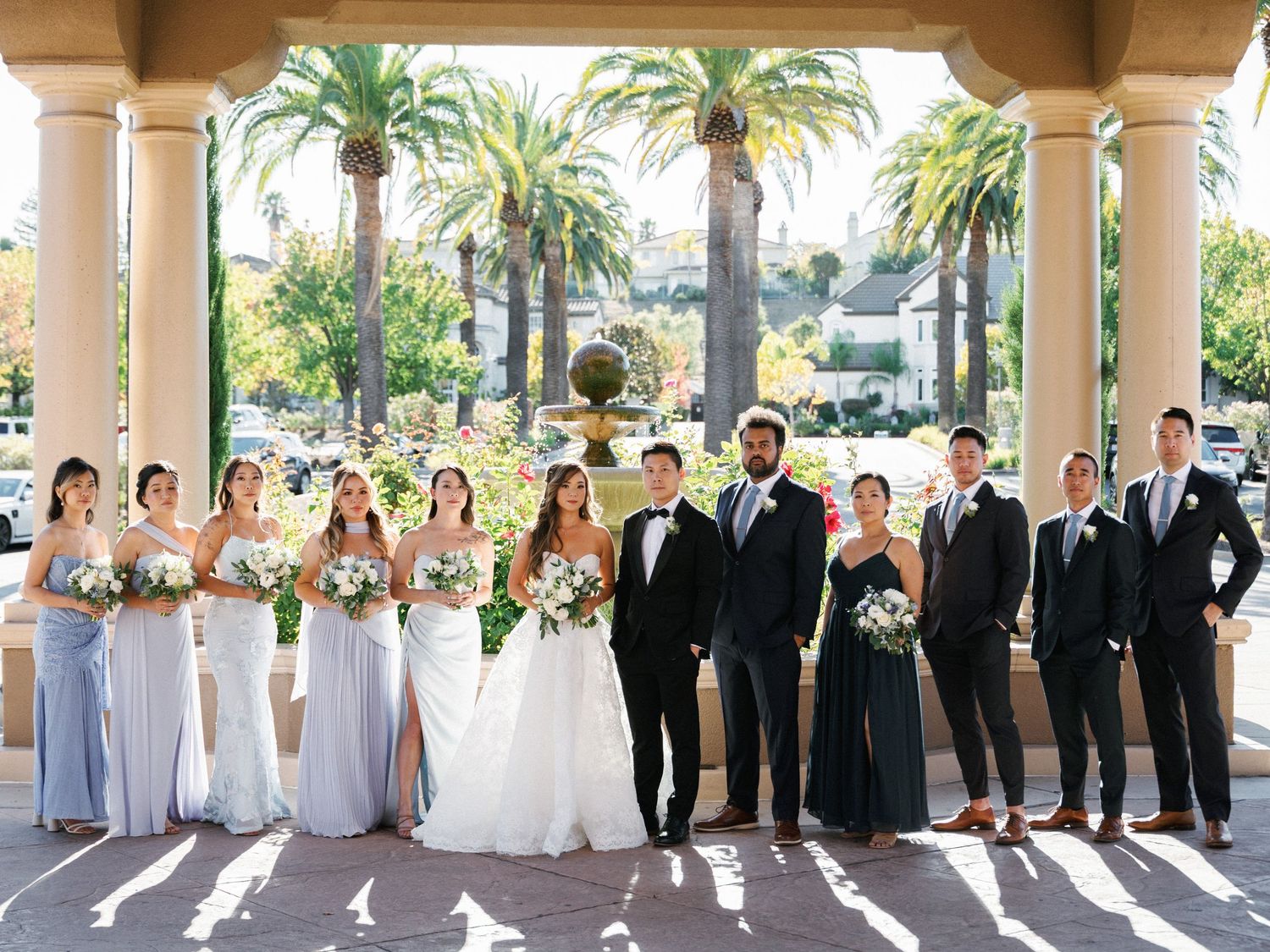 Wedding party in formal attire poses under Mediterranean-style archway with palm trees in background.