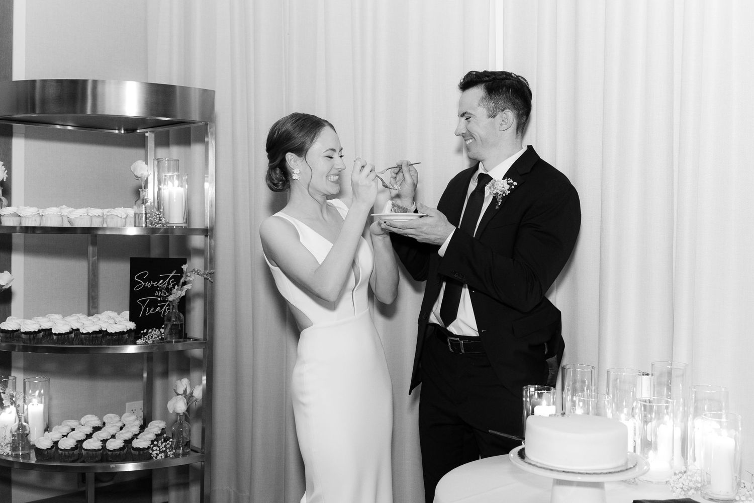 A couple in formal wedding attire share a playful moment while feeding each other cake at their reception.