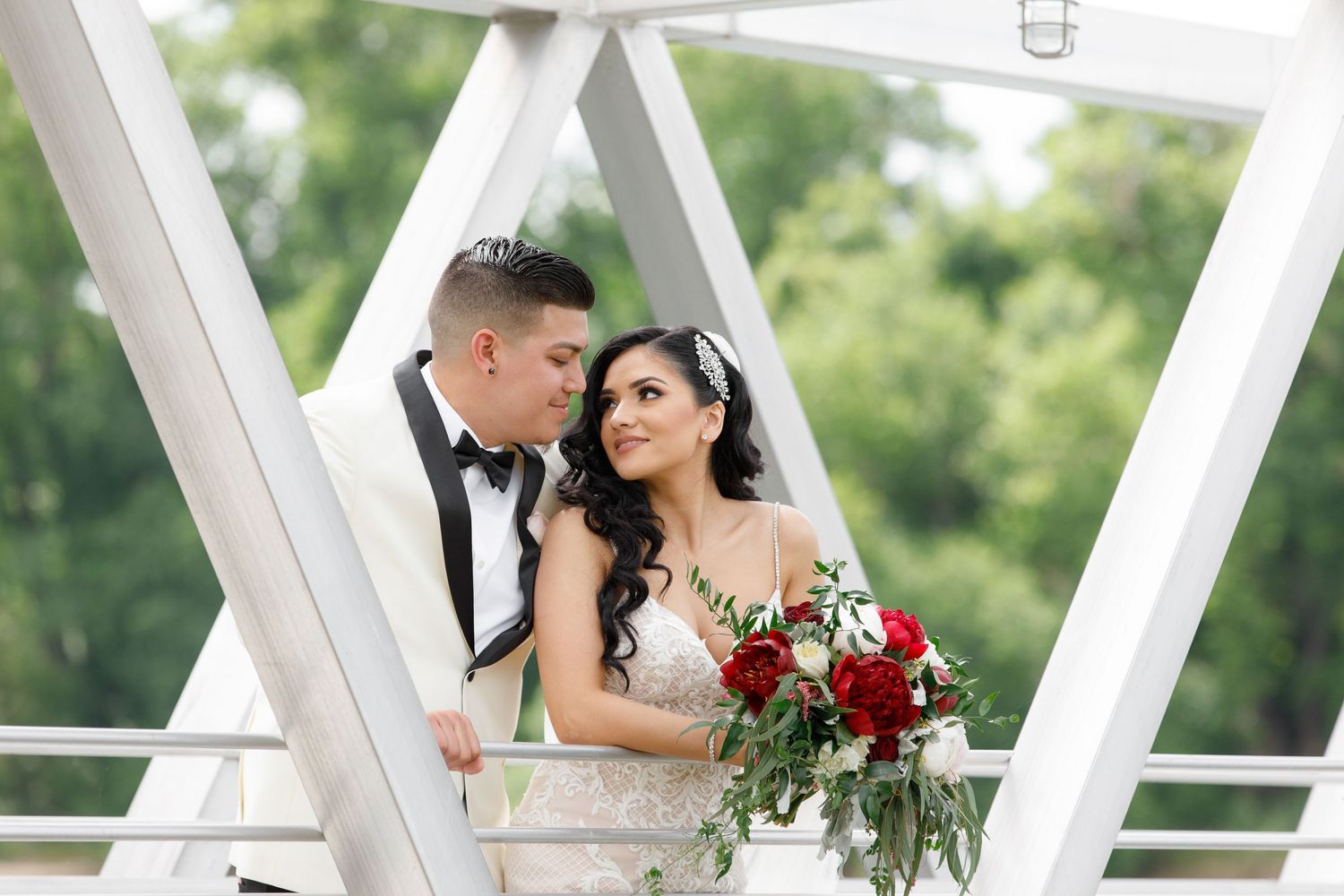 A wedding couple shares romantic moments on a white architectural bridge surrounded by lush greenery and red roses.