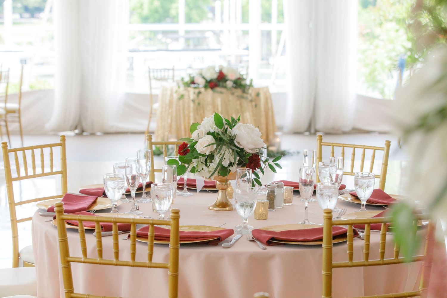 Elegant wedding reception tables with gold chiavari chairs and blush floral centerpieces in a bright white venue.