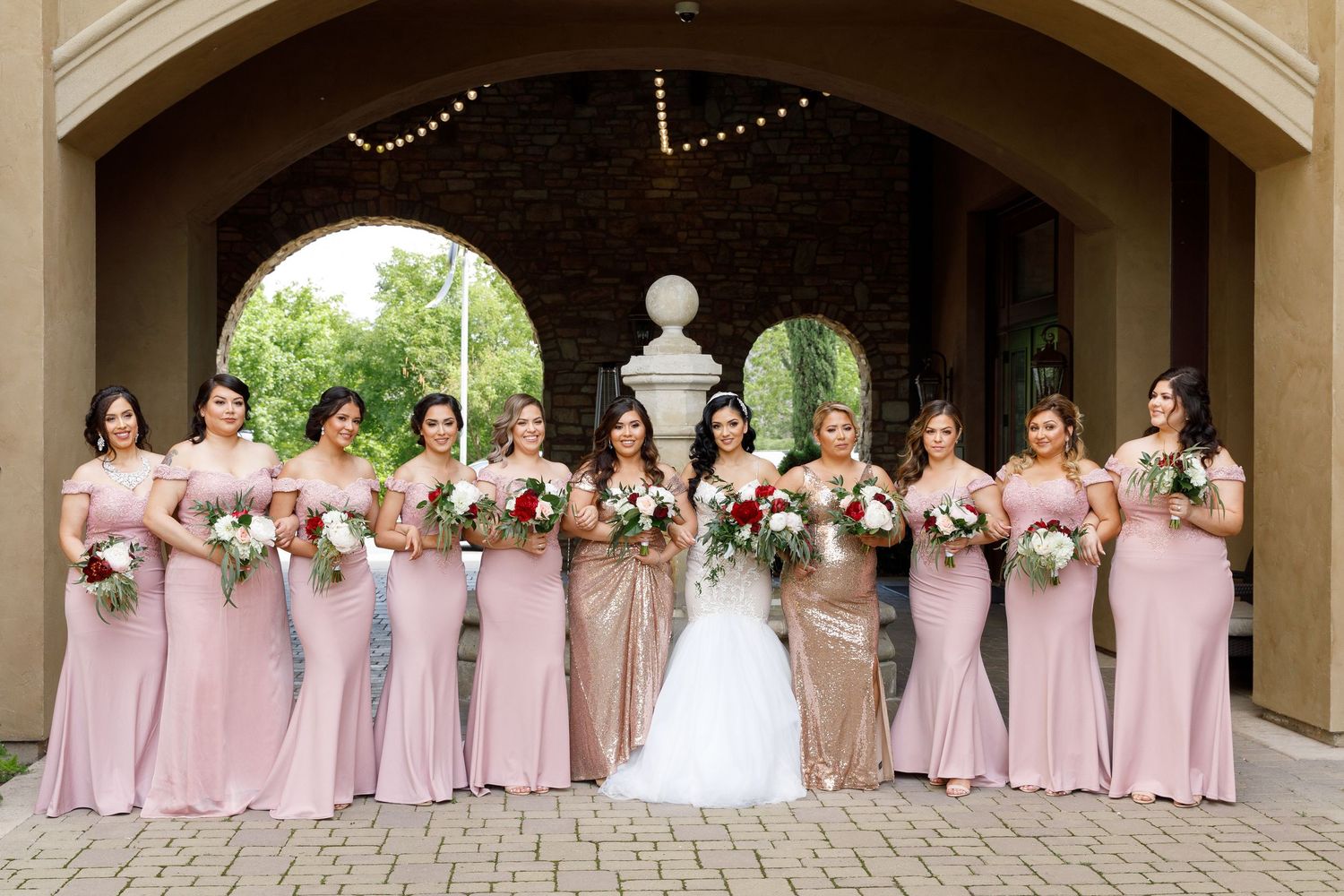 A wedding party poses in formal attire with bridesmaids wearing matching pink dresses in an elegant outdoor setting.