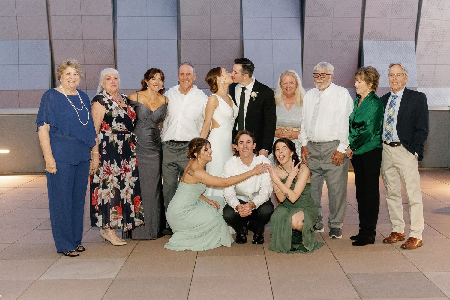 Wedding party poses together for a group photo against a modern wall backdrop at an indoor venue.