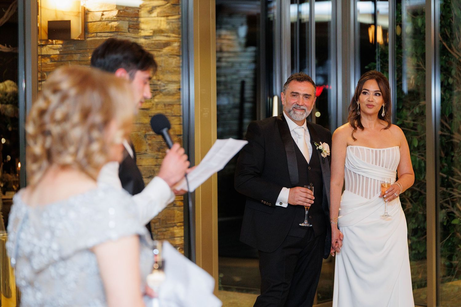 Wedding guests listen attentively during a speech at an elegant indoor reception venue with glass doors.