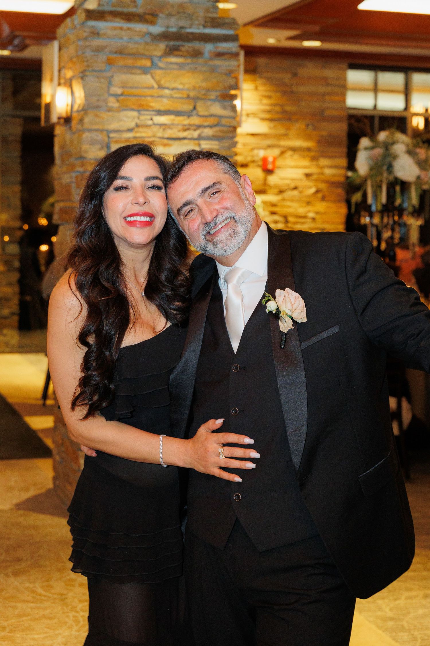 Couple in formal black attire sharing affectionate moments at an elegant venue with stone walls and warm lighting.