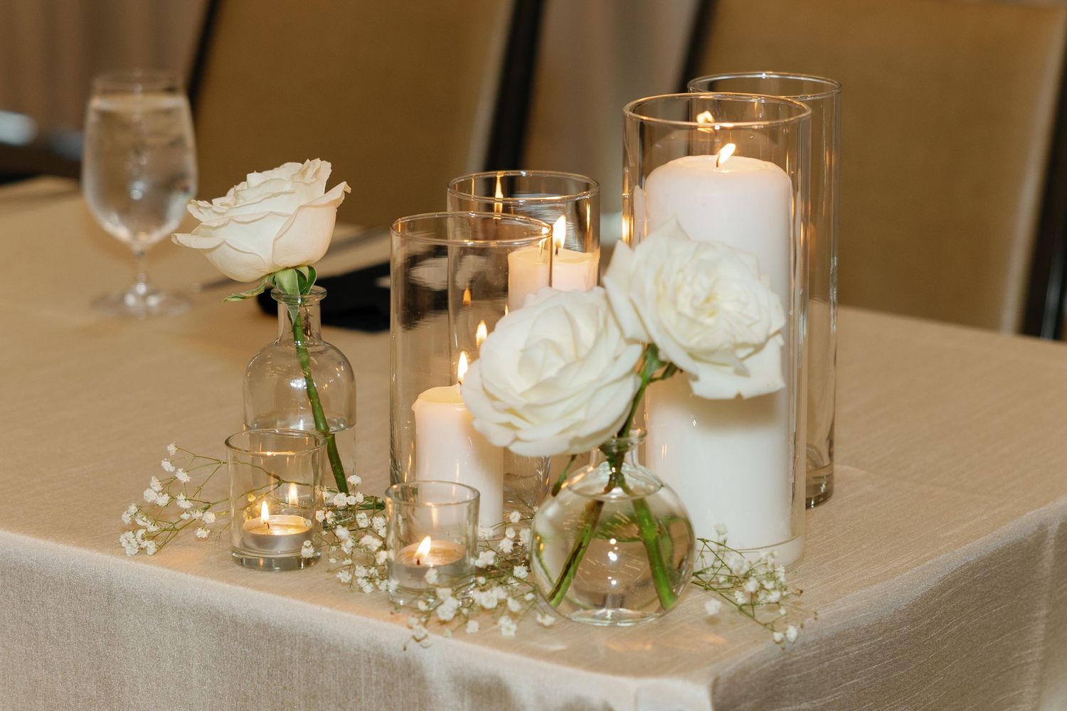 White roses and glowing candles create an elegant wedding reception centerpiece on a light-colored tablecloth.