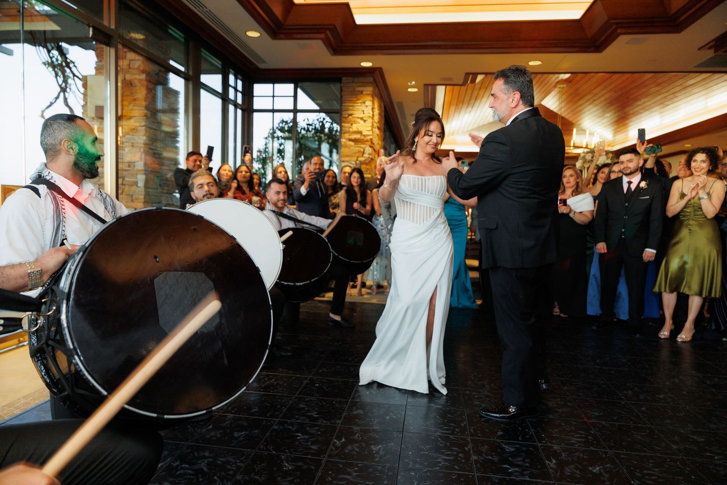 Wedding guests dance together on a dark wood dance floor during an evening reception celebration.