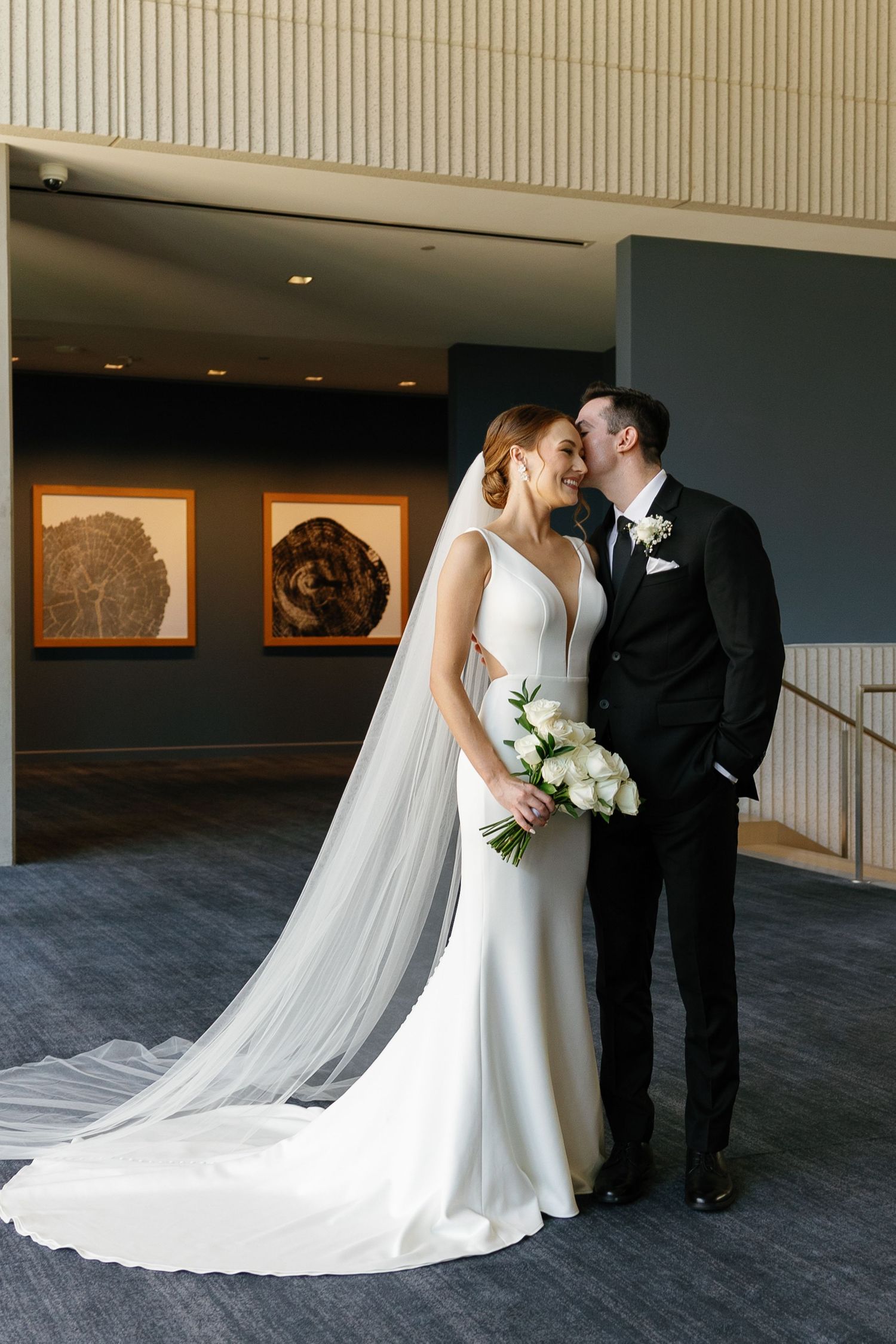 Elegant bride in sleek white gown with long veil shares intimate moment with groom in black suit at modern venue.