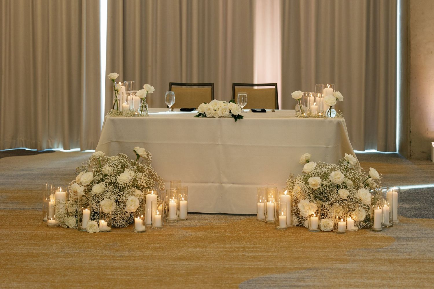 White floral arrangements and glowing candles create an elegant wedding reception setup along a grand ballroom wall.