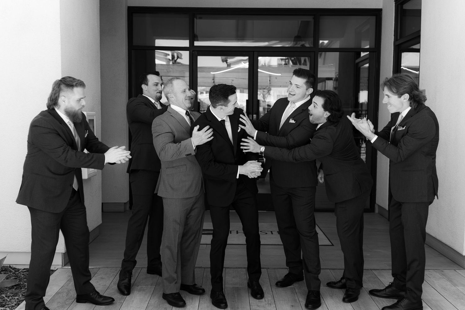 Black and white photo of groomsmen in suits laughing and drinking together before a wedding ceremony.