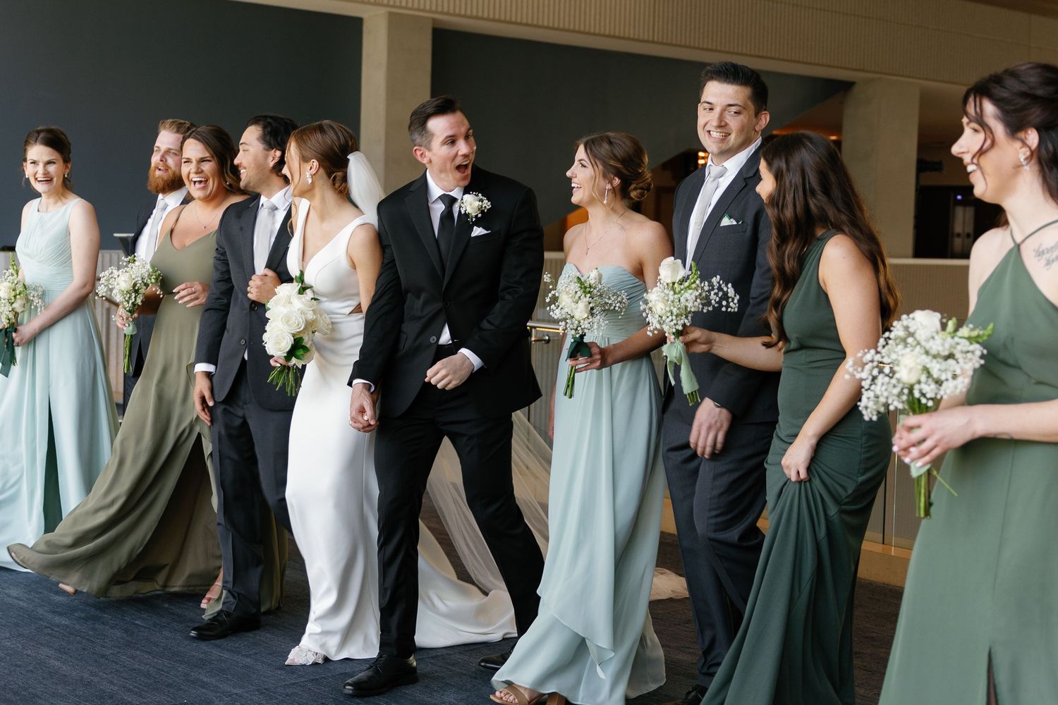 Wedding party members laugh and dance together during a reception celebration in a bright indoor venue.