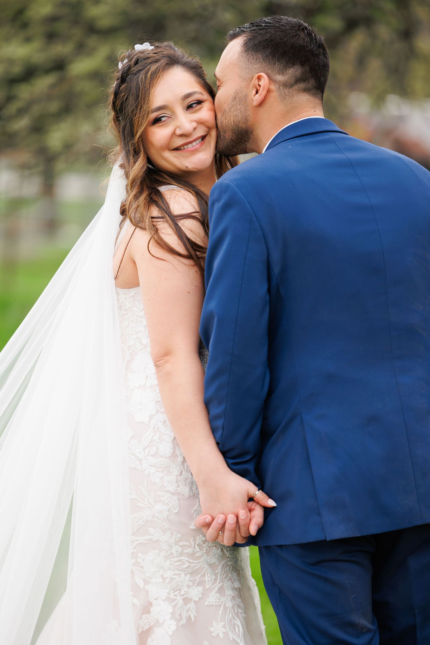 A bride in a white lace dress and groom in navy suit share intimate moments during their outdoor wedding photoshoot.