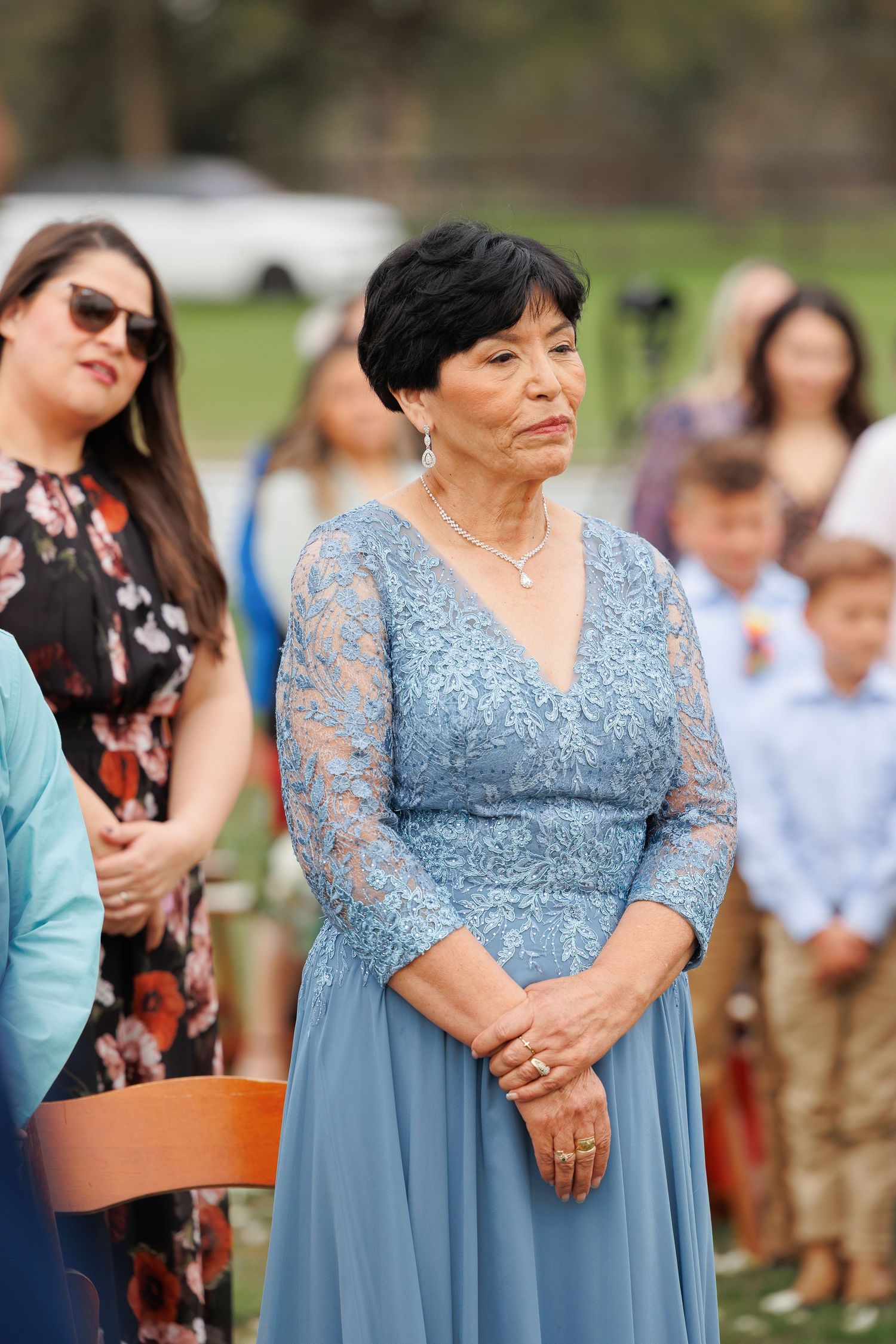 A group of wedding guests in light blue and floral dresses stand together during an outdoor celebration.
