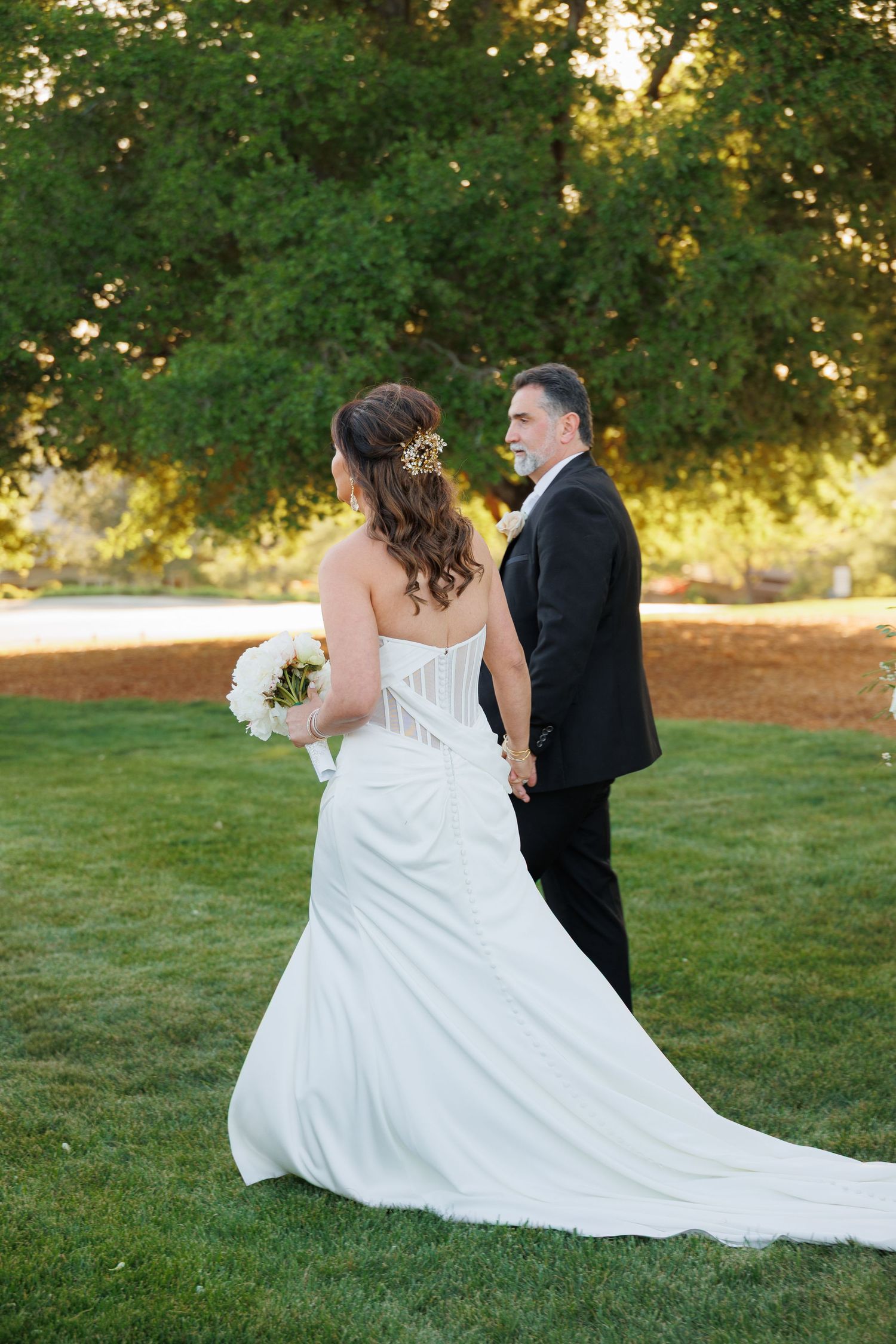 A couple in wedding attire sits together on lush green grass during sunset, with the bride's white dress flowing behind them.