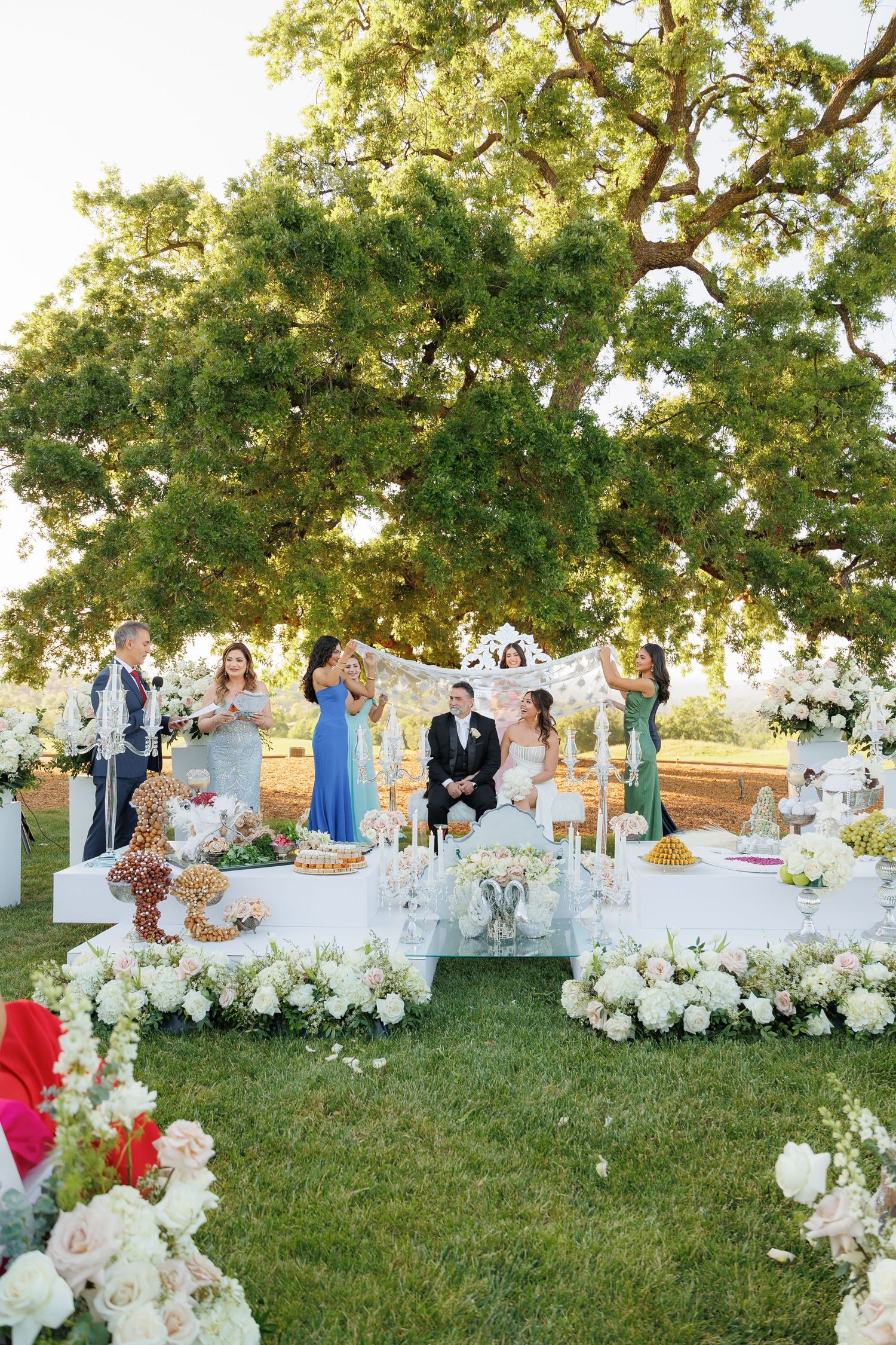 Wedding guests gather on a grassy hillside for an elegant outdoor reception with white floral arrangements and seating.