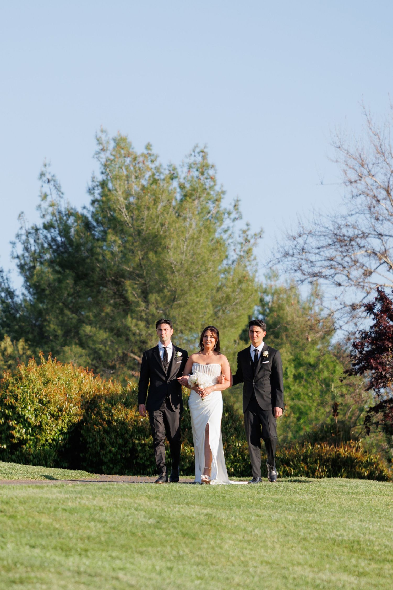 Wedding party walks together across a grassy hilltop on a sunny autumn day with trees in the background.