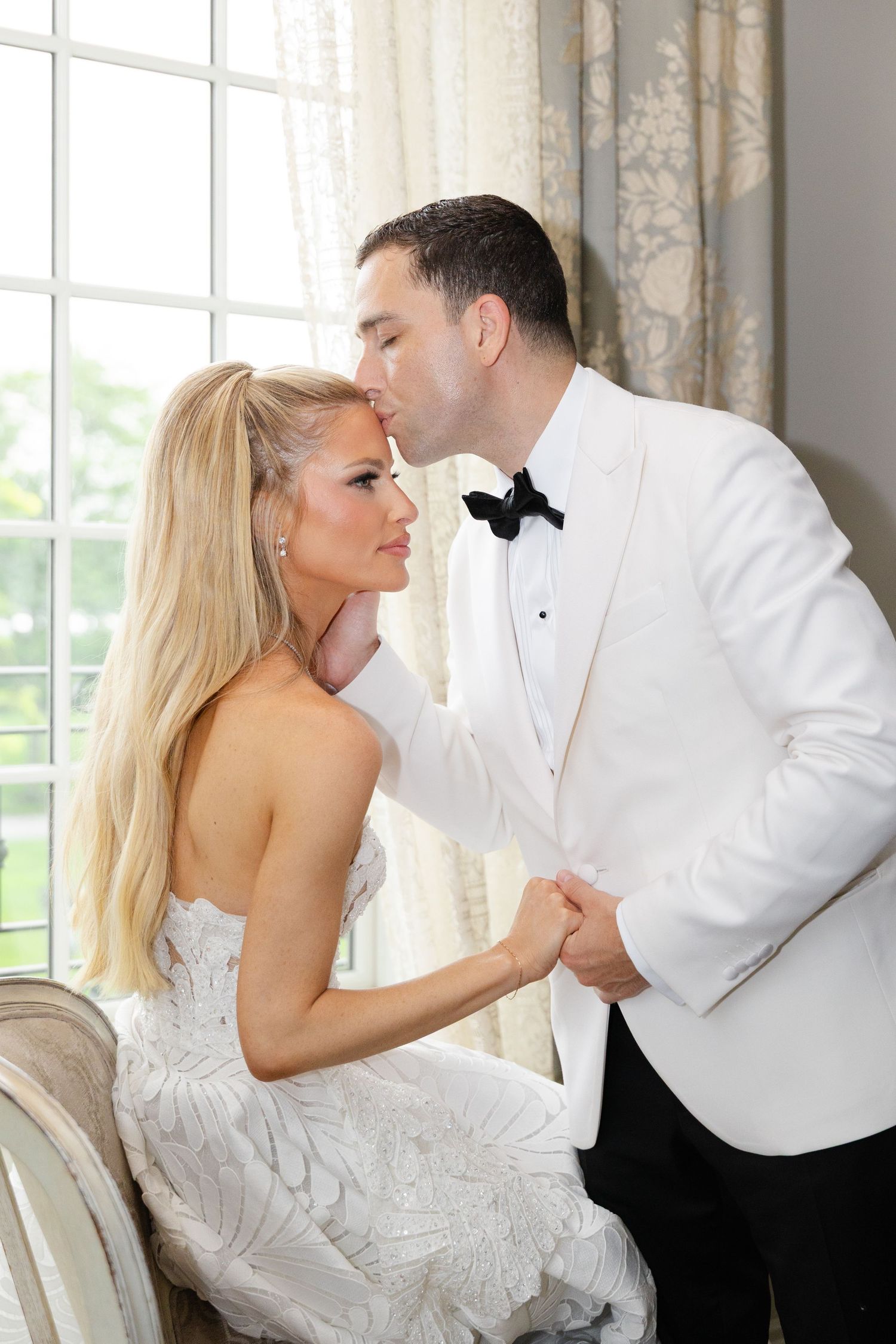 A couple in formal wedding attire share an intimate dance moment in an elegant room with window light.