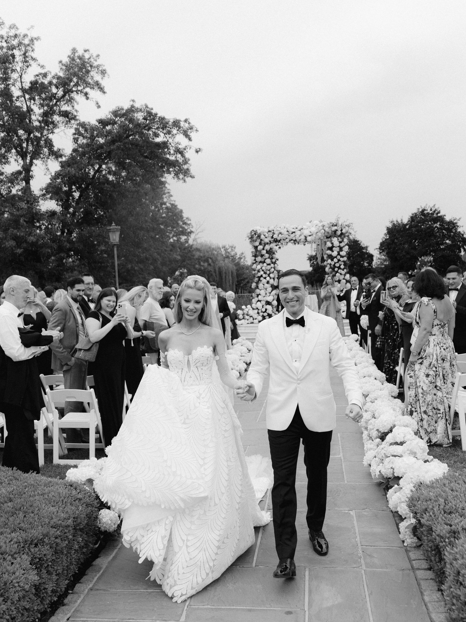 Wedding party members walk down an outdoor aisle lined with white flowers and greenery at an elegant garden ceremony.