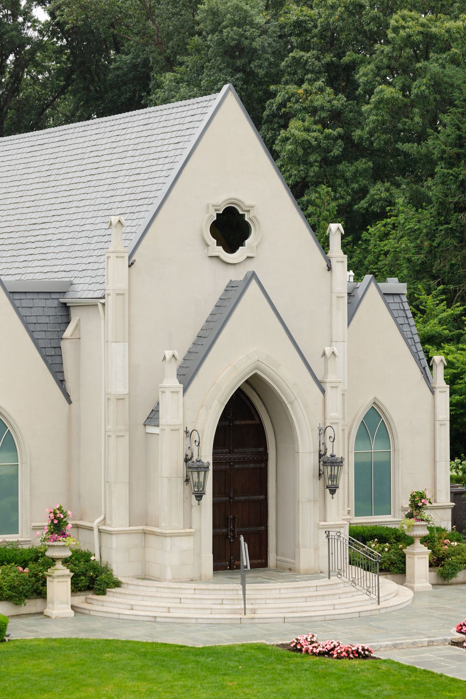 A white Gothic-style church chapel with pointed arched doorways and windows surrounded by lush green landscaping.