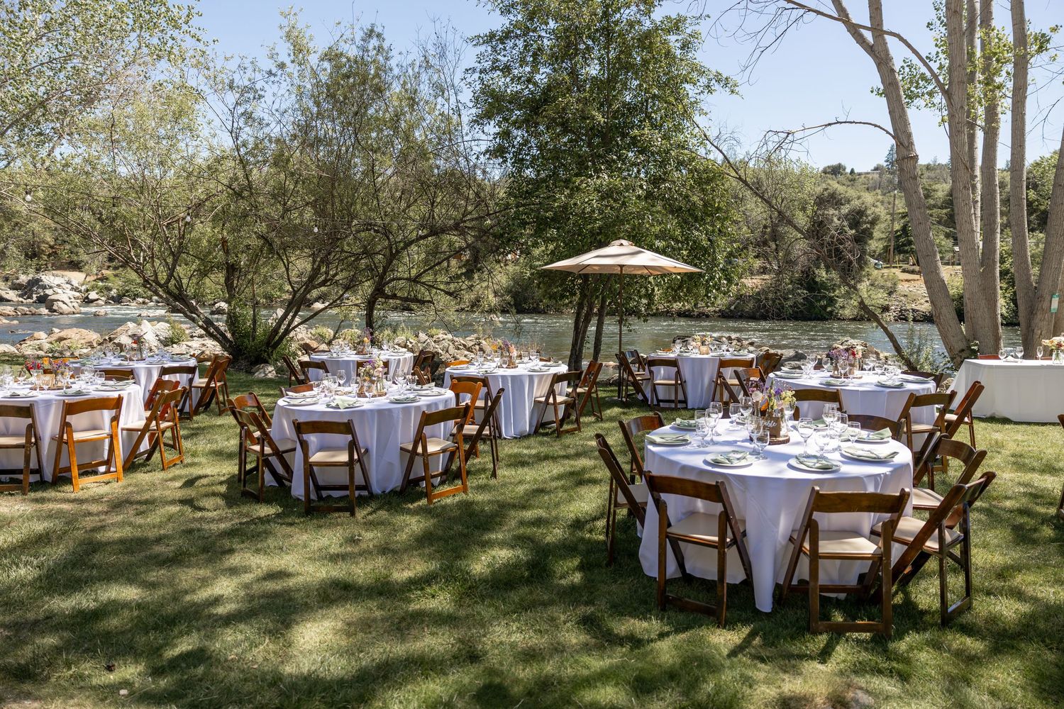Elegant outdoor wedding reception tables set under shady trees with white tablecloths and wooden chairs.
