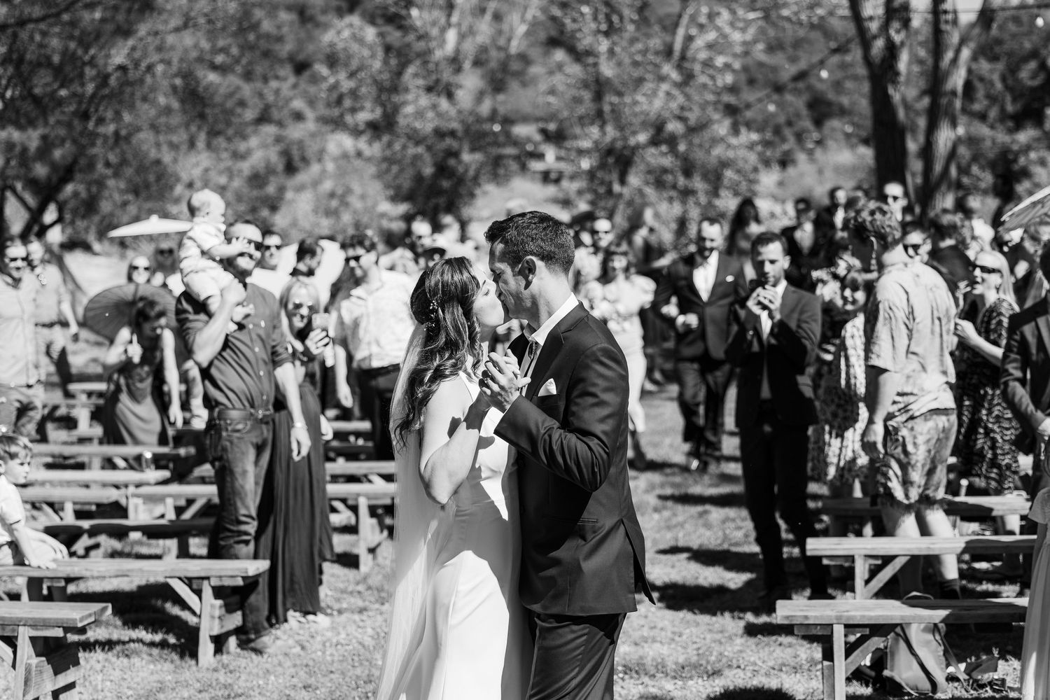 Wedding guests gather outdoors in a park setting for a casual celebration with tables and chairs arranged under trees.