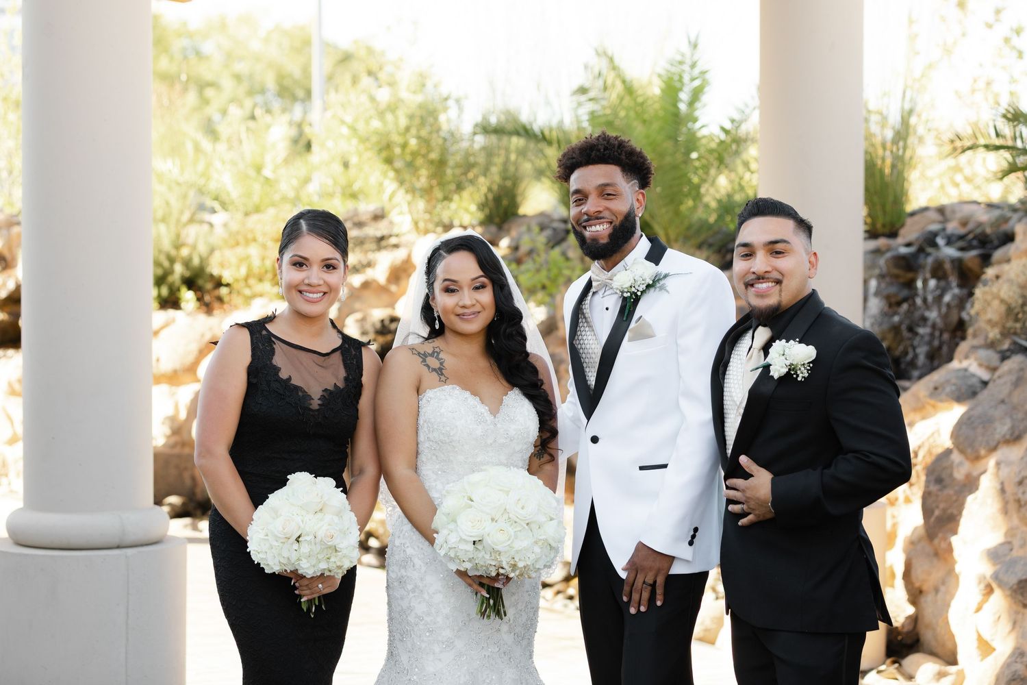 Wedding party in formal attire poses together between white columns during an elegant outdoor celebration.