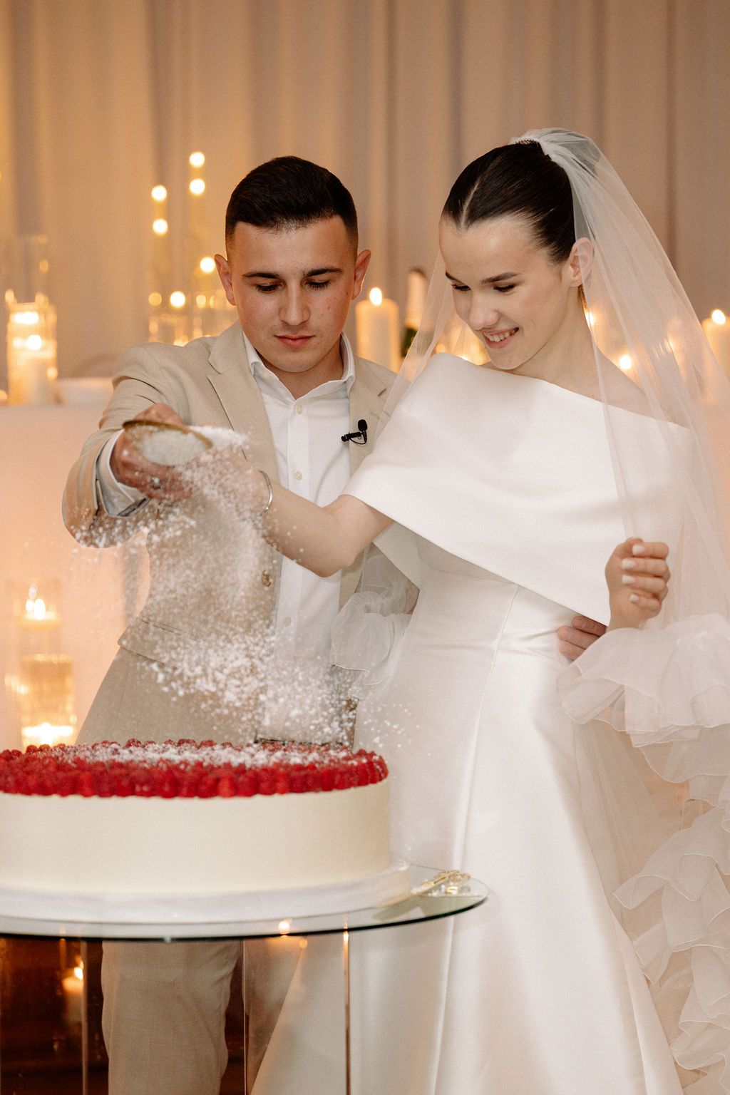 Wedding couple cuts into their elegant white tiered cake with red rose decorations in a candlelit reception venue.