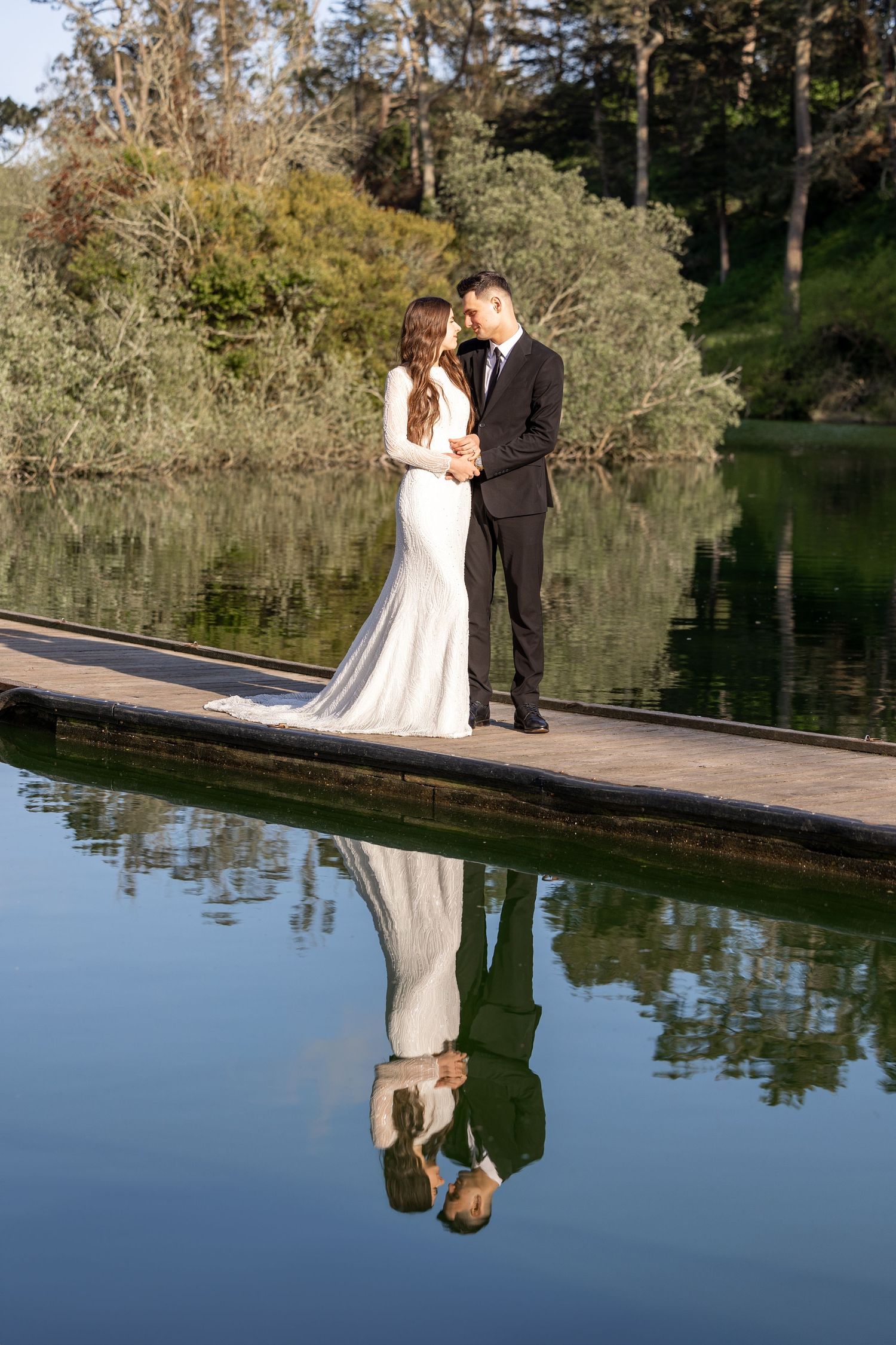 Wedding couple reflected in still lake water with trees in background.