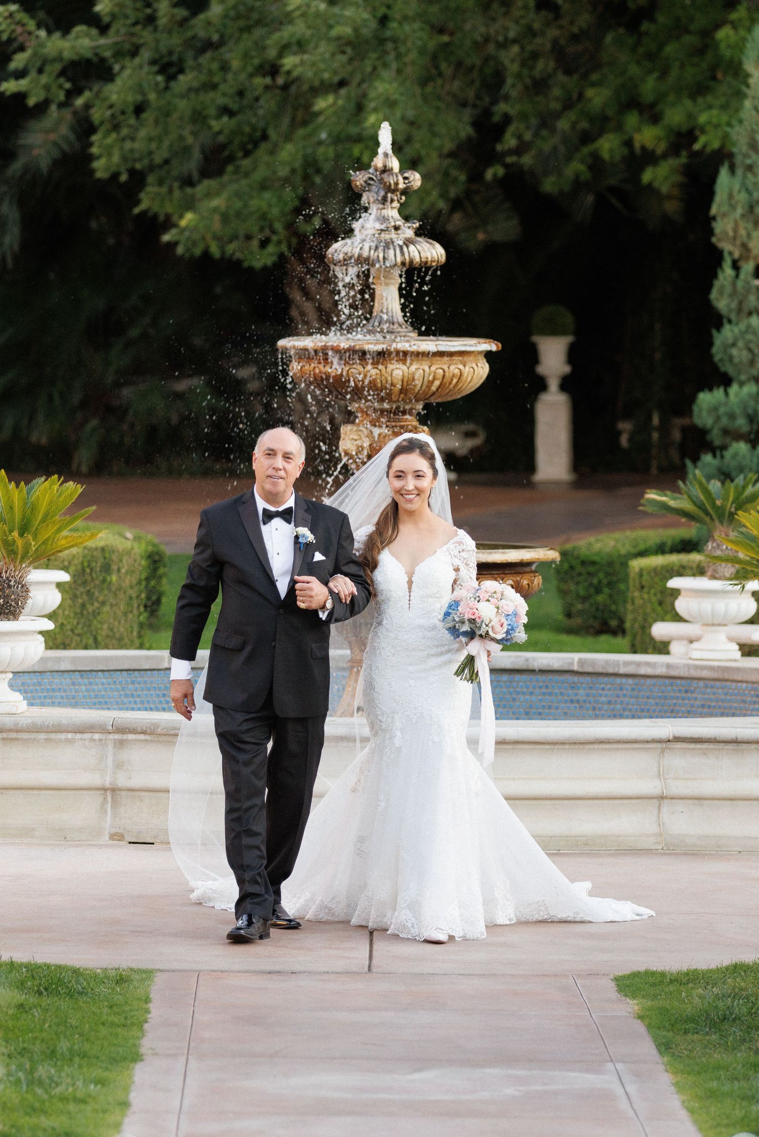 Elegant wedding couple walking along stone path by ornate garden fountain in formal attire.