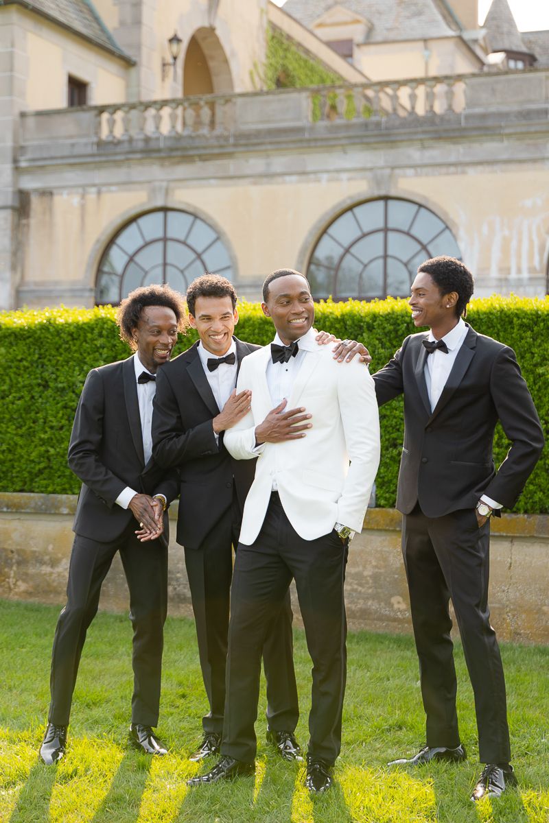 Groomsmen in black tuxedos share laughs on the lawn of an elegant estate.
