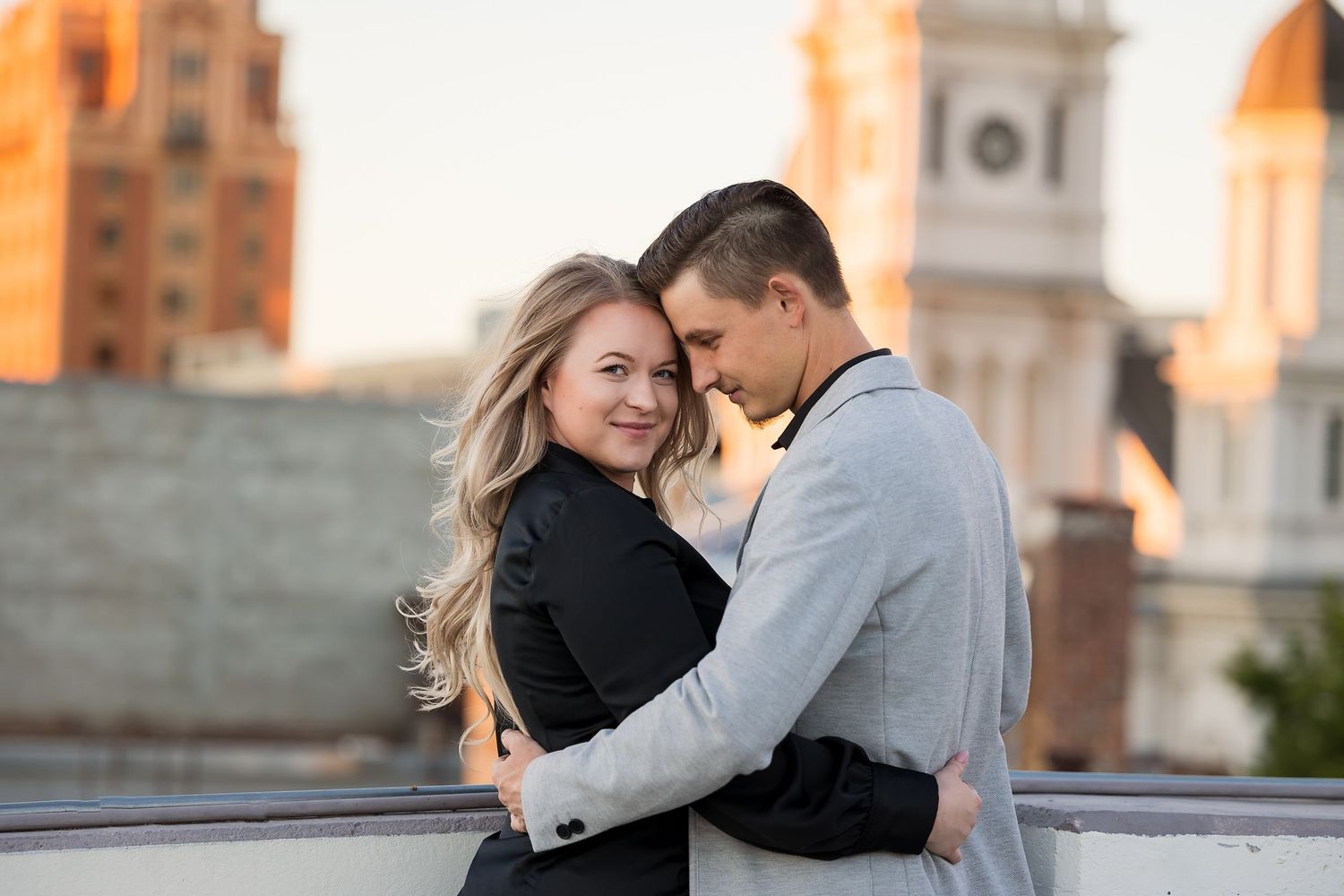 A couple embraces and smiles on a rooftop at sunset with historic buildings in the background.