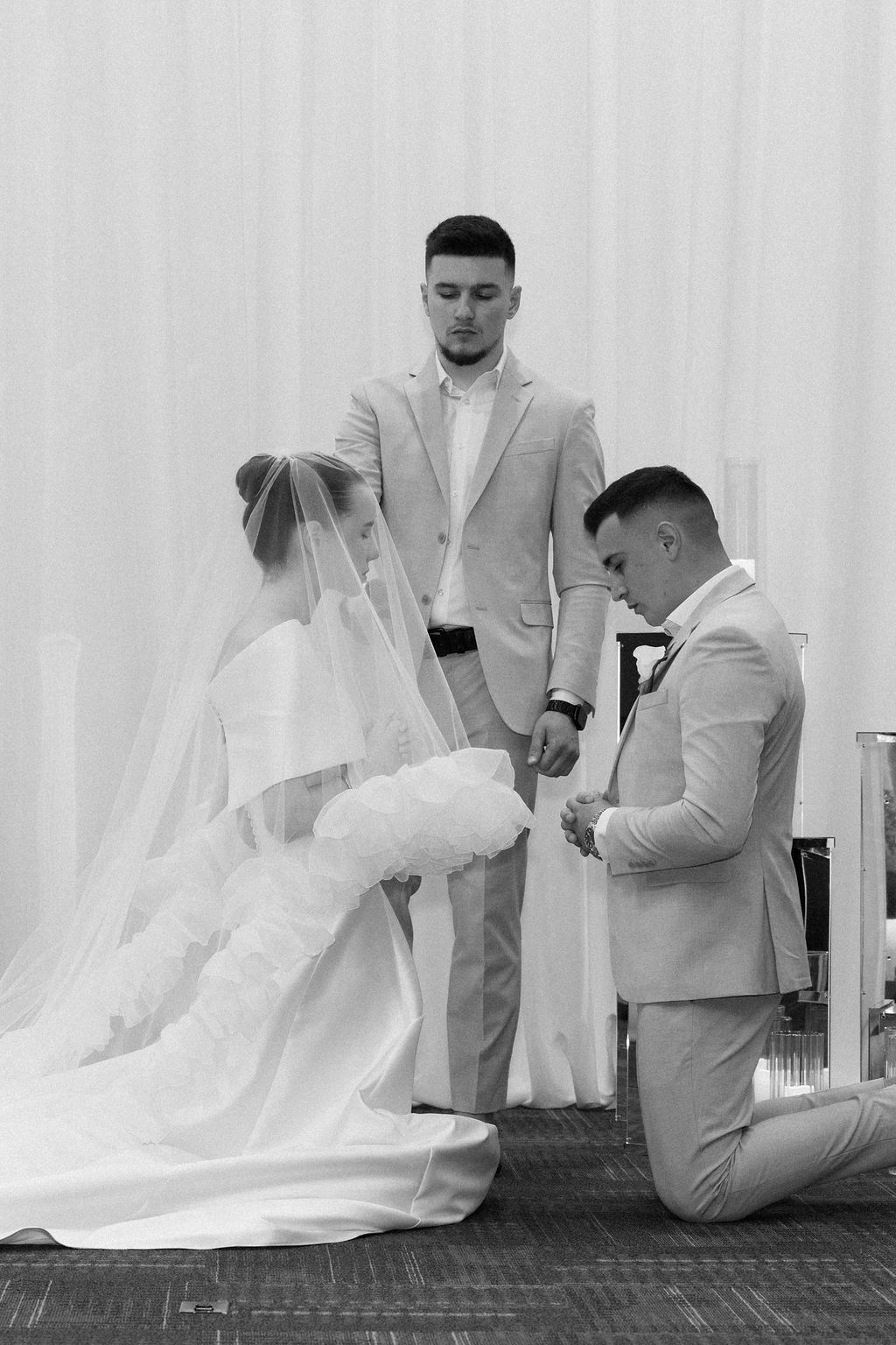 A black and white wedding ceremony photo showing a bride in a flowing gown with a veil and two men in light suits.